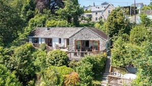 Une maison en pierre dotée d’un balcon, entourée d’une végétation luxuriante et bordée d’autres maisons au loin.