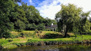A serene riverside scene with a quaint house surrounded by lush greenery and trees. - Penwaun, Newport - Three Bedroom Cottage, Sleeps 8 (Nevern)