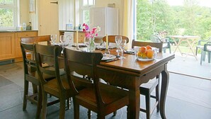 A dining area with a wooden table set for four, surrounded by chairs. There is a window with a view of greenery outside. - Penwaun, Newport - Three Bedroom Cottage, Sleeps 8 (Nevern)