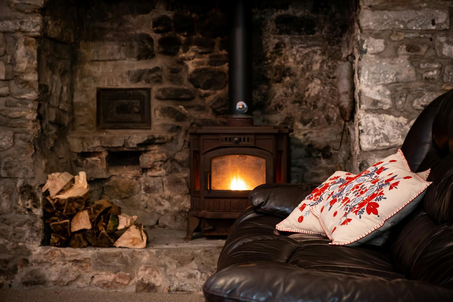 A stone fireplace with a wood-burning stove, a leather sofa with a decorative pillow, and a pile of firewood.