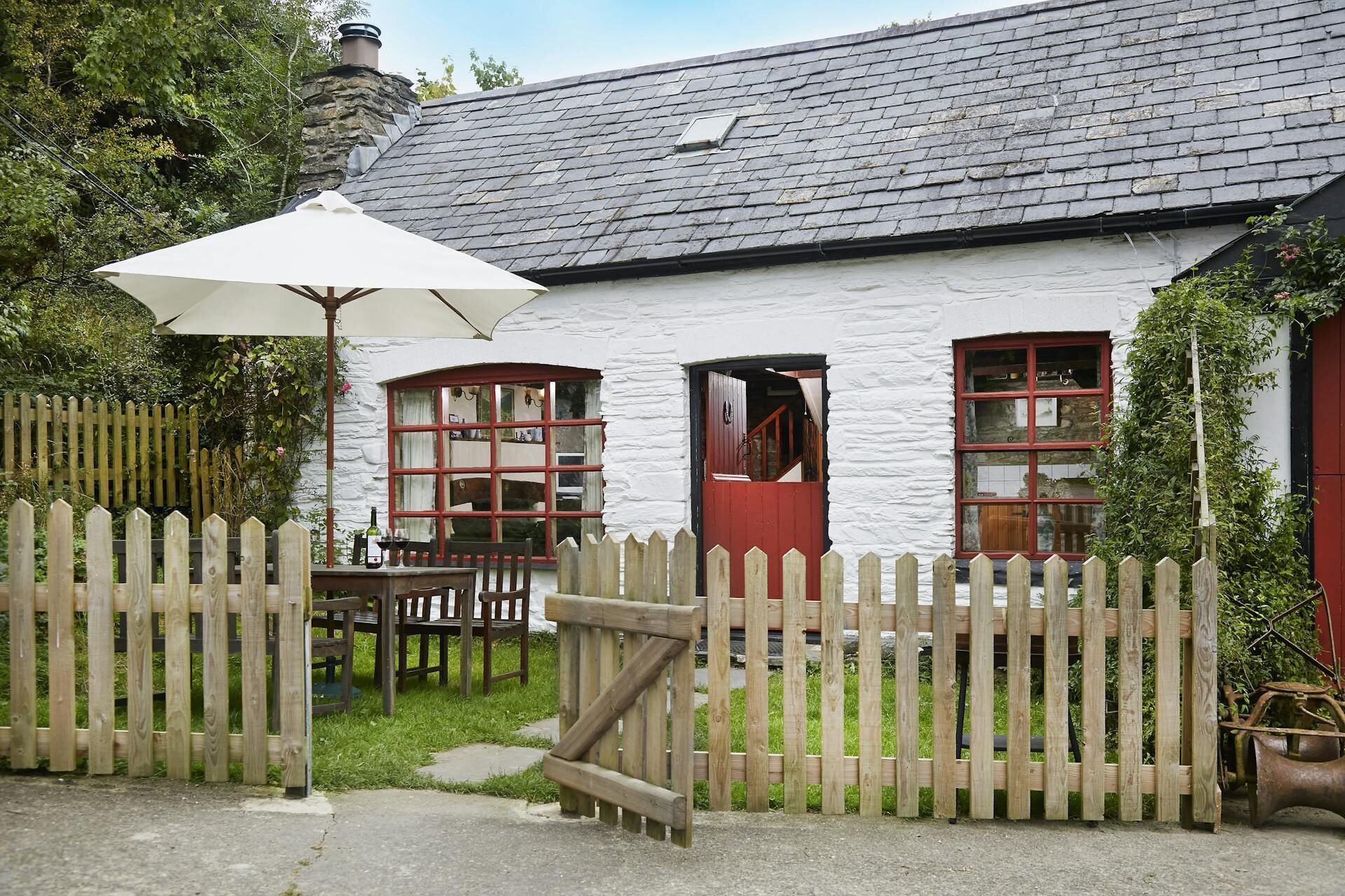A white building with a stone chimney, wooden fence, and a table with chairs outside.