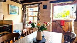 A rustic dining area with a wooden table, chairs, a vase of flowers, a clock, and a window with a view of greenery. - Y Beudy, Llangrannog (Llangrannog)