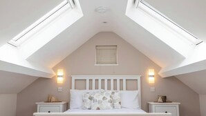 Une chambre avec un plafond en pente, des fenêtres de toit, un lit avec une tête de lit blanche, deux tables de chevet et des lampes fixées au mur.