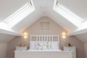A bedroom with a sloped ceiling, skylights, a bed with a white headboard, two bedside tables, and wall-mounted lights.