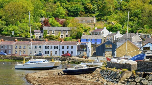 Une ville côtière où des bateaux sont amarrés près du rivage, des bâtiments aux façades en pierre et, en arrière-plan, une végétation luxuriante.