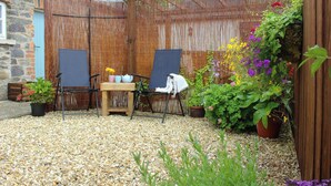 A garden with a wooden pergola, two folding chairs, a small table with a teapot and cups, and various potted plants and flowers.