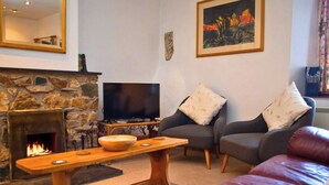 A living room with a stone fireplace, a flat-screen TV, a wooden coffee table, two grey sofas with patterned cushions, and a framed painting on the wall.