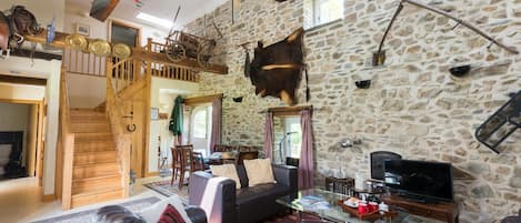 A rustic interior with a stone wall, wooden staircase, leather sofas, a dining area, and a mounted animal hide.
