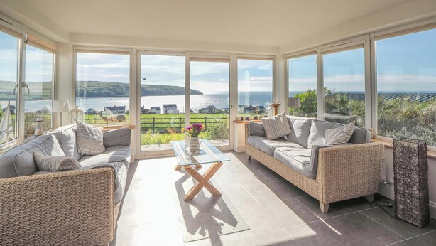 A spacious room with large windows, a glass coffee table, two sofas, and a view of a body of water. - Pentir, Gwbert - Four Bedroom Cottage, Sleeps 7 (Gwbert)