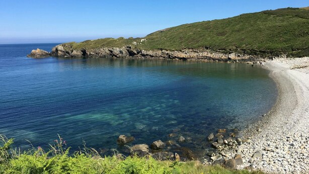 A coastal landscape with clear blue waters, a pebble beach, and a grassy hill.