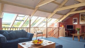 A spacious loft with a high ceiling, wooden beams, a blue sofa, a wooden coffee table with fruit, and a TV in the background.