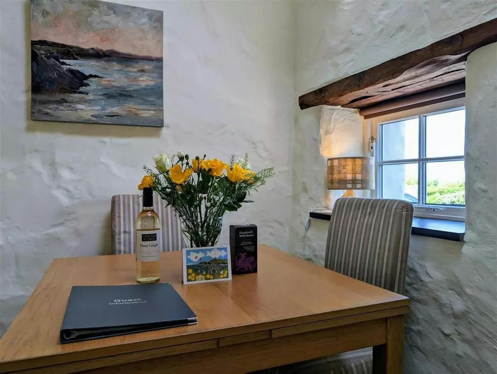 Un tavolo in legno con un laptop, una bottiglia di vino, un vaso di fiori gialli e una cornice con una foto.