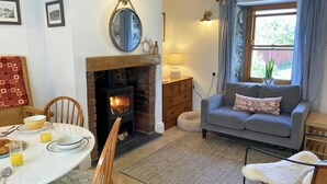 A cozy living room with a fireplace, a dining table with breakfast items, a sofa with a cushion, and a window with curtains. - Y Bwthyn, Newport - Two Bedroom Cottage, Sleeps 4 (Newport)