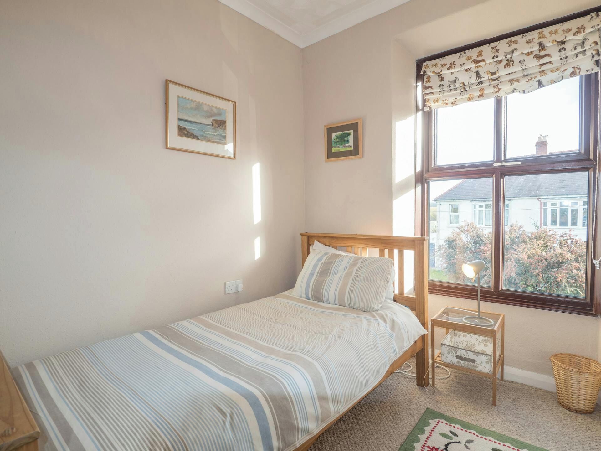 Un dormitorio con una cama individual, una mesita de noche, una lámpara, un ventanal con vistas a casas y dos cuadros en la pared.