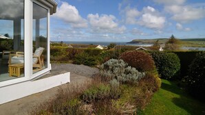 Una casa moderna con un invernadero de vidrio, un jardín con diversos arbustos y vistas al mar y las colinas al fondo.