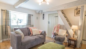 Un salon chaleureux avec un canapé gris, un pouf vert, une chaise en bois et une petite table avec une lampe. On aperçoit un escalier au fond.