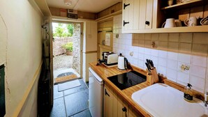 Une cuisine compacte équipée d’un lave-vaisselle encastré, d’un évier et d’une cuisinière. Une petite fenêtre se trouve au-dessus de l’évier, et une porte donne accès à un espace jardin.
