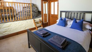 A bedroom with a bed, pillows, a bedside table, a lamp, a wooden staircase, and a stone wall. - Parrog Stores (Newport)