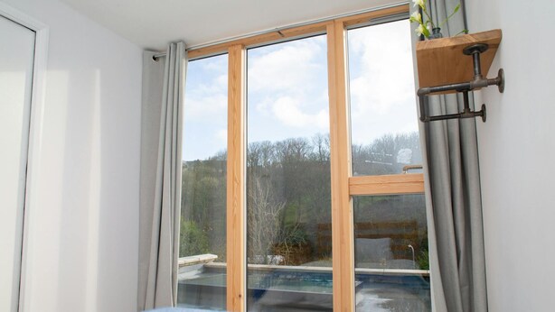 A bedroom with a large window, a bed with a blue and white blanket, and a wooden shelf with a vase. - 5 Seabird Close, Little Haven (Little Haven)