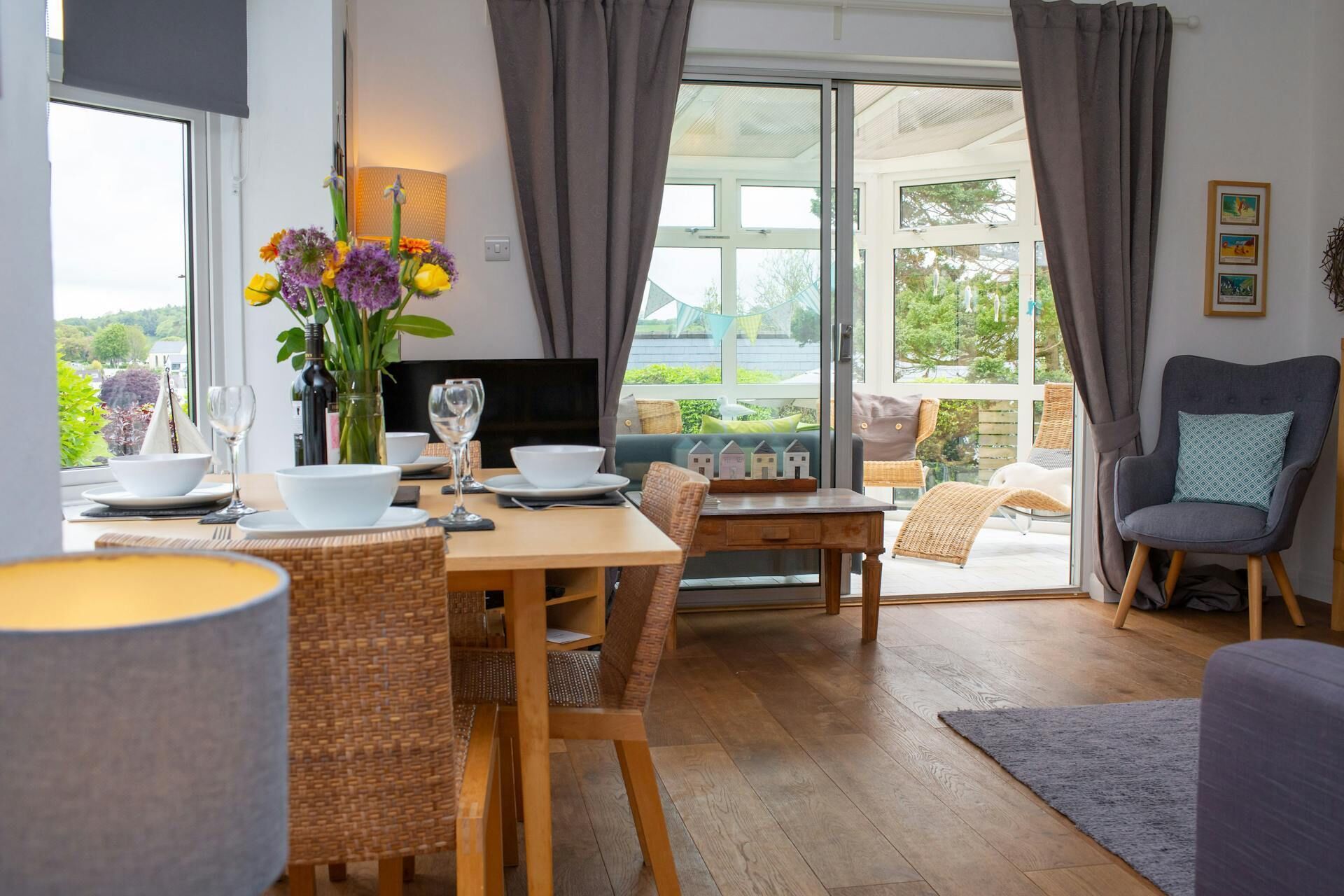 A dining area with a table set for two, a vase with flowers, and a view of the outdoors through a glass door.