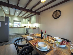 A dining area with a wooden table set for a meal, a kitchen in the background, and a clock on the wall. - Bryndalis, Aberaeron (Dihewyd)