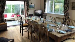 Un coin repas avec une table en bois dressée pour six personnes, entourée de chaises, d’un téléviseur et d’un canapé agrémenté de coussins rouges.