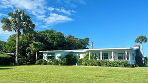 A single-story house with a white exterior, surrounded by a well-maintained lawn and various trees, including palm trees.
