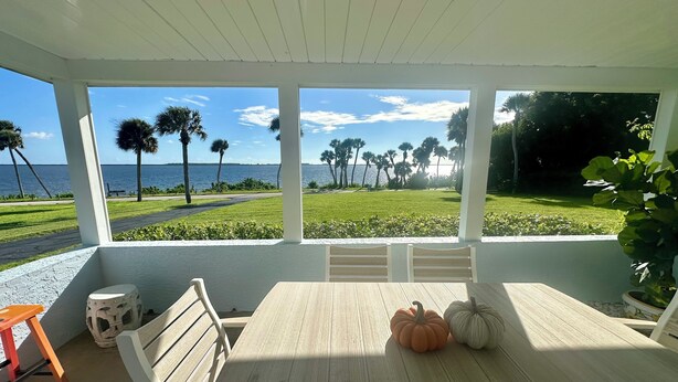 A covered patio with a table and chairs, two pumpkins, and a view of a grassy area with palm trees and a body of water.