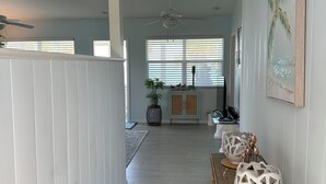 A hallway with a white wall, a ceiling fan, a large window with blinds, a patterned rug, and a wicker table with decorative vases.