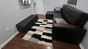 A modern living room with a sectional sofa, a black ottoman, a checkered rug, and a television mounted on the wall. - Grandiosely Pleasant Suite 2 (Hempstead)