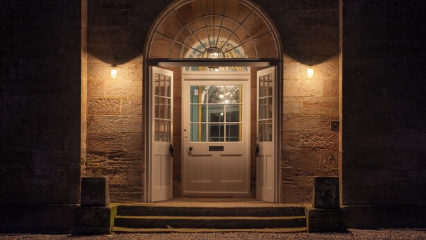 Entrada de un edificio con un amplio arco, una puerta blanca y dos luces fijadas a la pared.