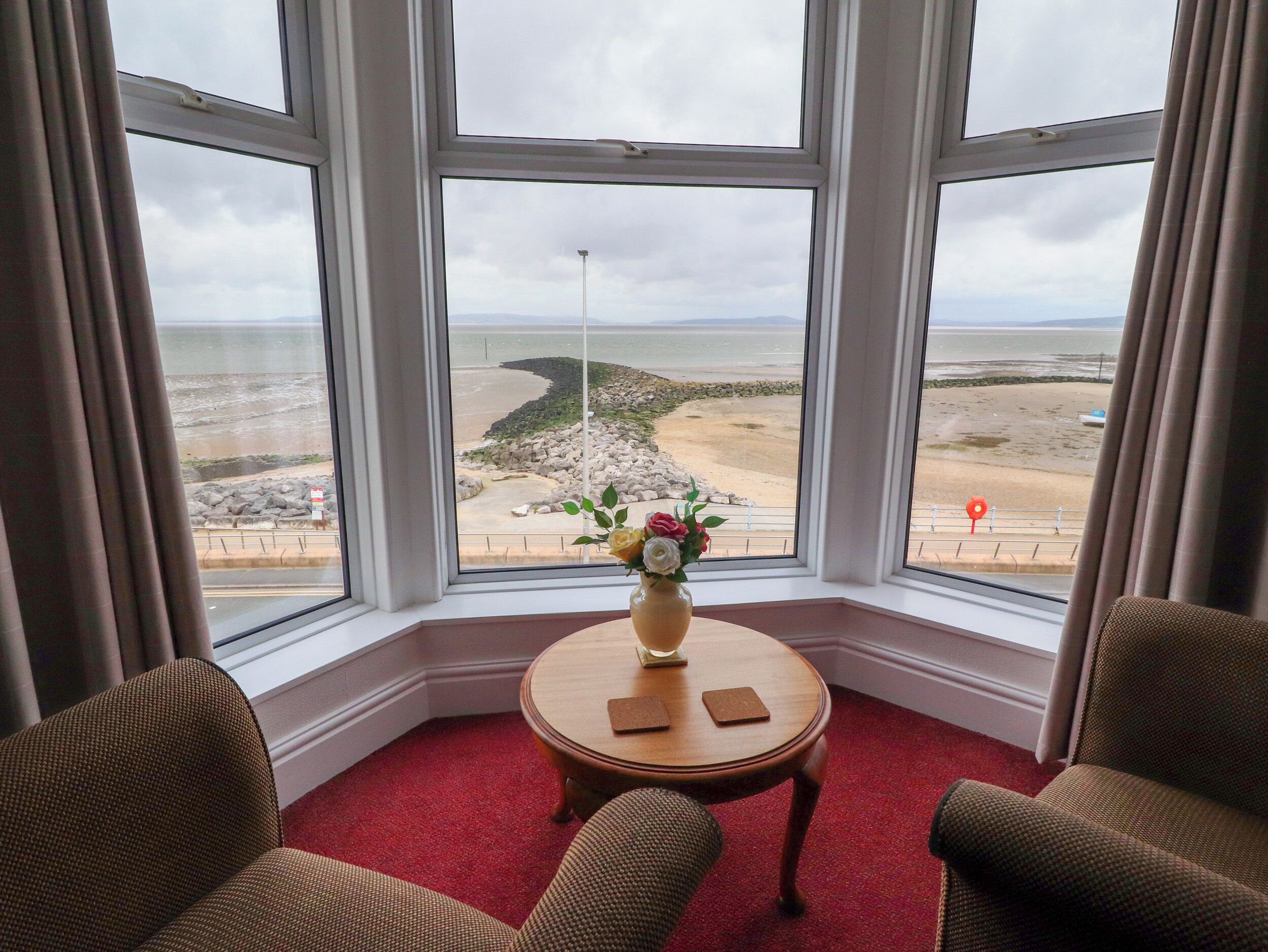 Une chambre d’angle dotée d’une grande baie vitrée donnant sur une plage. On y trouve une petite table ronde avec un vase de fleurs et deux sous-verres. Deux fauteuils rembourrés sont placés devant la table.