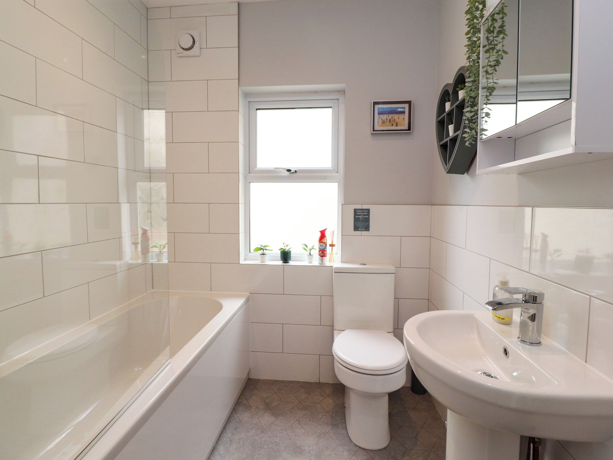 A bathroom with white tiles, a bathtub, a toilet, a sink, and a window with plants.