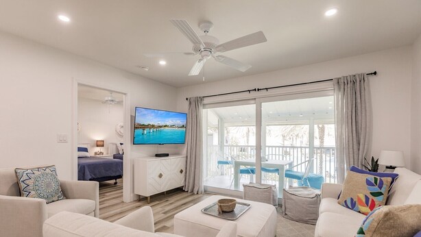 A modern living room with a flat-screen TV, white furniture, a ceiling fan, and a balcony with a view of the sea.