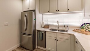 A modern kitchen with a stainless steel refrigerator, a sink with a window above, and light-colored cabinetry.