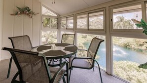 Ein sonnendurchfluteter Raum mit einem runden Tisch und vier Stühlen, großen Fenstern mit Blick auf ein Gewässer und Grünflächen sowie einem Deckenventilator.