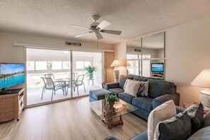 A modern living room with a sofa, coffee table, and a television. There is a balcony with a view of the sea.