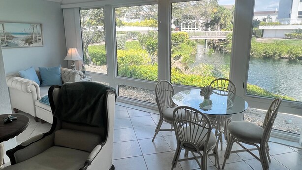 A room with a dining table and chairs, a sofa with cushions, a ceiling fan, and large windows overlooking a water body.