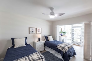 Une chambre avec deux lits, un ventilateur de plafond, une table de chevet avec une lampe et une fenêtre avec des stores.