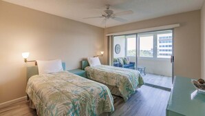 Habitación de hotel con dos camas, ventilador de techo, balcón con vistas y escritorio con lámpara.