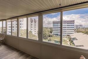 A room with large windows, a chair, and a view of a modern building and greenery.