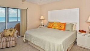 Un dormitorio con cama, mesita de noche, lámpara, silla y vista al océano.