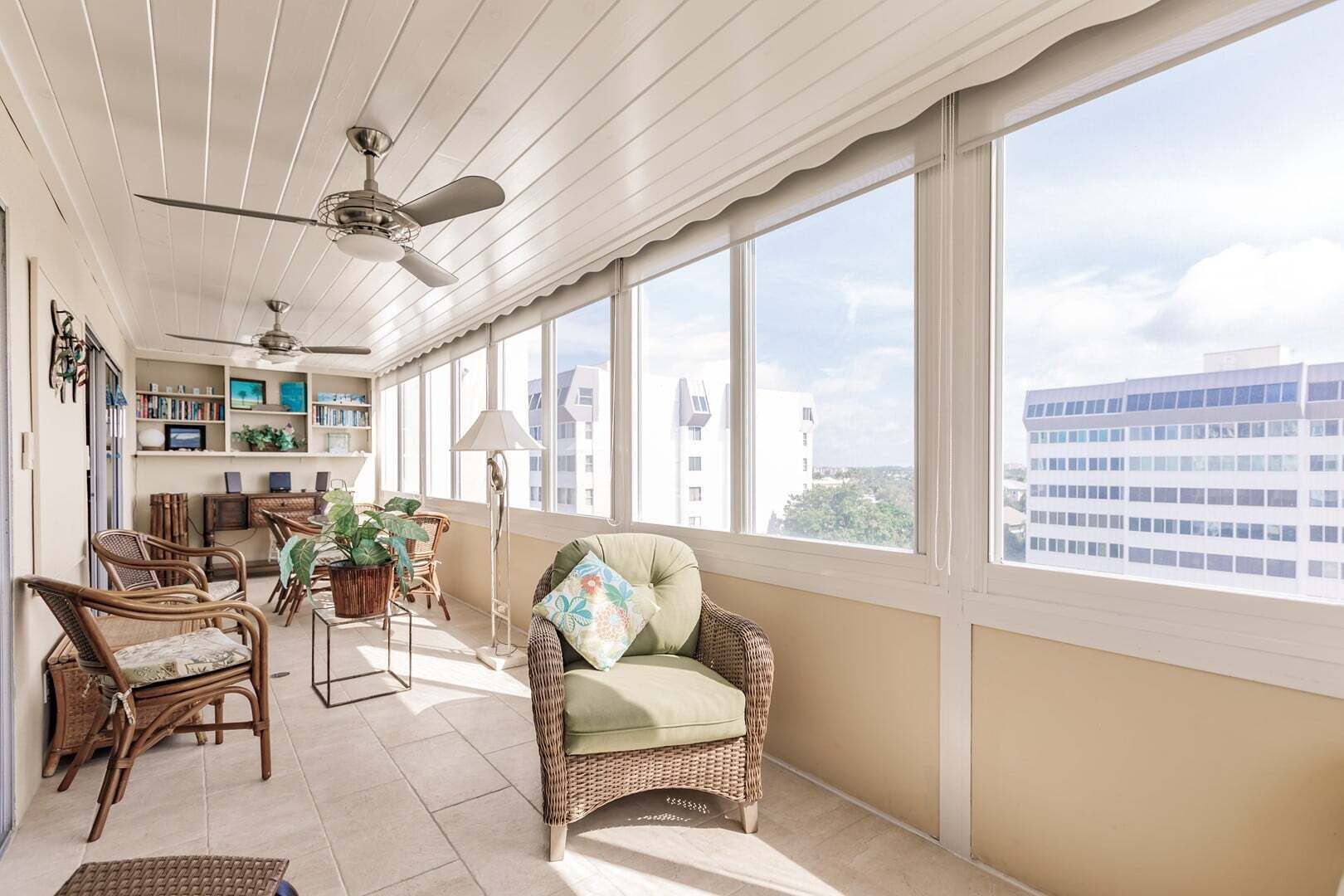 A sunlit room with wicker furniture, a ceiling fan, and a view of buildings outside.