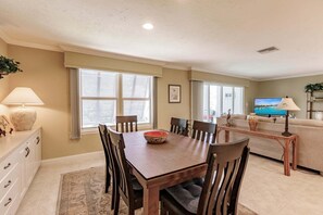 A dining area with a wooden table and chairs, a television in the background, and a sofa in the living room.