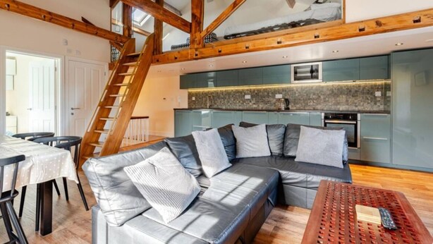 A modern loft-style living space with exposed wooden beams, a built-in kitchen, and a cozy seating area. - Sedbergh Penthouse - Kingbed - Tv - Wifi (Sedbergh)