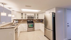 A modern kitchen with white cabinets, stainless steel appliances, a built-in microwave, oven, and refrigerator. There is a skylight above the stove and a small island with a sink and countertop.