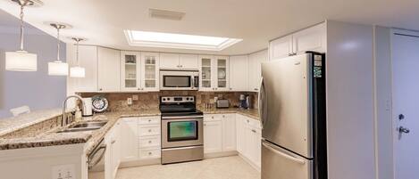 Cocina moderna con gabinetes blancos, electrodomésticos de acero inoxidable, microondas integrado, horno y refrigerador. Hay un tragaluz sobre la estufa y una pequeña isla con pileta y encimera.