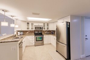 Cocina moderna con gabinetes blancos, electrodomésticos de acero inoxidable, microondas integrado, horno y refrigerador. Hay un tragaluz sobre la estufa y una pequeña isla con pileta y encimera.