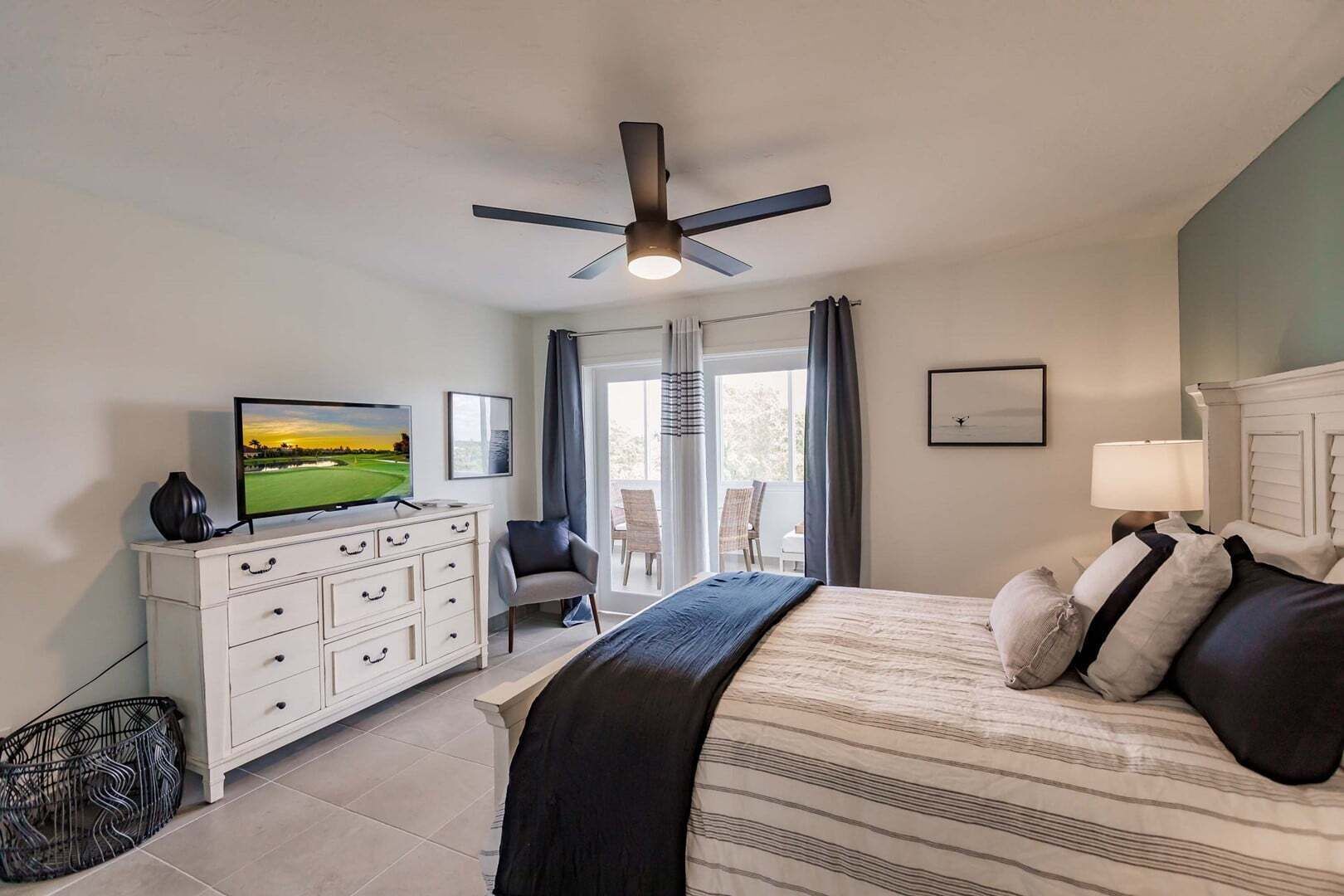 A bedroom with a bed, a TV, a ceiling fan, a dresser, and a view of the outdoors.
