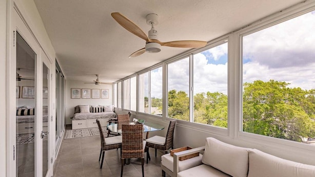 A sunlit room with a ceiling fan, a sofa, a dining table with chairs, and a view of trees through large windows. - 1125-407 - Premium Plus - Step into Coastal Paradise (Siesta Key)